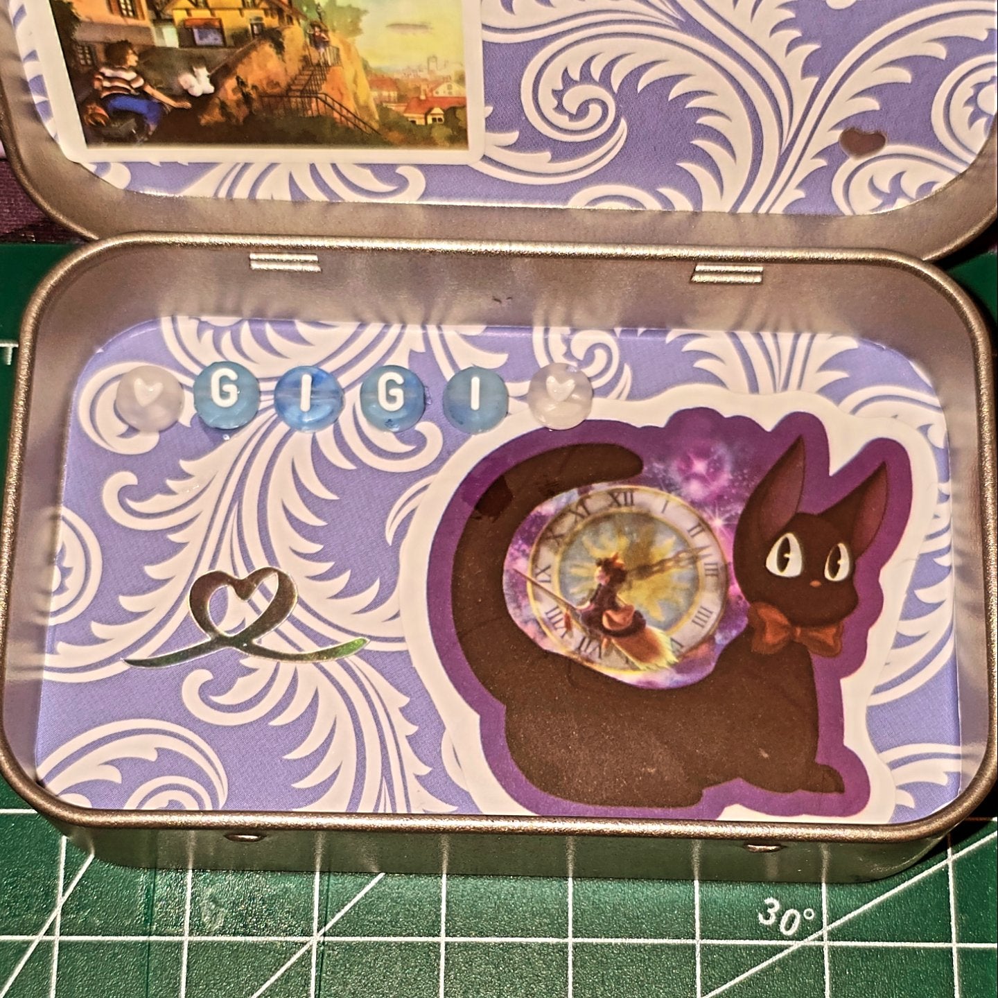Decorative tin with a cat and clock design on a purple background, placed on a cutting mat.